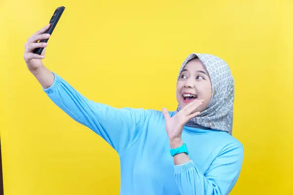 Tesettür ve mavi gömlekli neşeli Asyalı bir kadın el sallarken, gülümserken ve telefonuna bakarken selfie çekiyor..