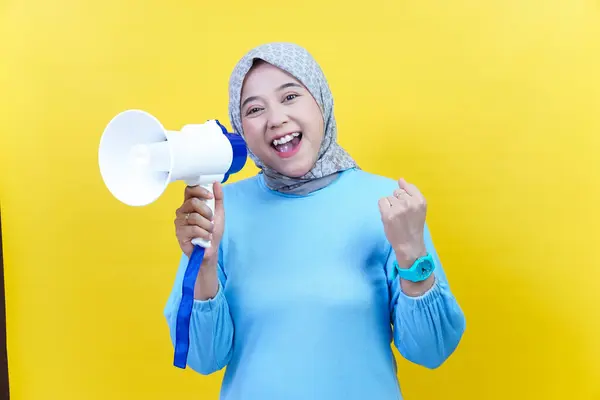 Tesettürlü mutlu bir Asyalı kadın bir megafon tutuyor ve sarı arka planda heyecanla yumruğunu kaldırıyor..