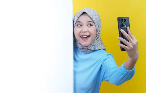 Beyaz duvarın arkasında tesettürlü neşeli bir kadın akıllı telefonuyla selfie çekerken sarı bir arka plana bakıyor..