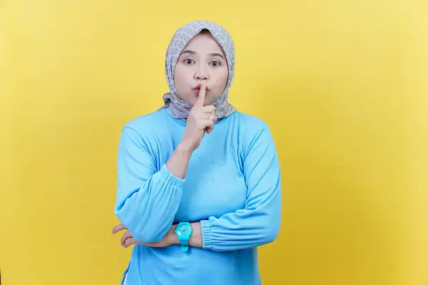 Tesettürlü ve mavi elbiseli Asyalı bir kadın parmaklarıyla dudaklarında sessizlik işareti yapıyor..