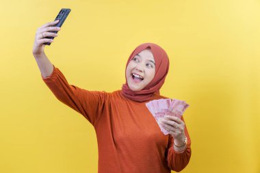 Tesettürlü mutlu bir Asyalı Müslüman kadın elinde para tutarken selfie çekiyor, finansal başarı ve neşeyi temsil ediyor..