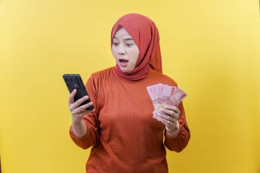 Elinde akıllı telefon tutarken elinde para tutan, beklenmedik finansal haberlere tepki veren şok olmuş Asyalı Müslüman bir kadın..