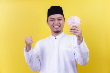 İslami kıyafetli neşeli bir Asyalı Rupiah 'ı elinde tutuyor ve sarıya karşı bir kutlama için elini kaldırıyor..