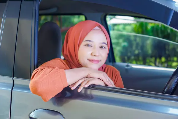 Asyalı, tesettürlü Müslüman bir kadın arabanın camına yaslanıyor, sıcak bir şekilde gülümsüyor, dışarıda huzurlu bir anın tadını çıkarıyor..