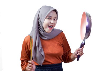 Tesettürlü neşeli Asyalı kadın elinde spatula ve tavayla yemek pişirme pozisyonunda gülümsüyor..
