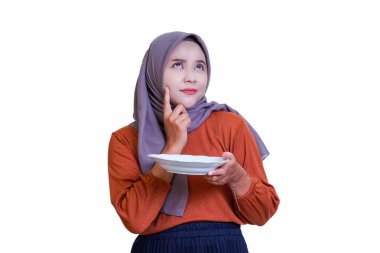 Gri tesettürlü ve turuncu tişörtlü Asyalı bir kadın bir tabak tutuyor ve düşünceli bir şekilde çenesine dokunuyor..