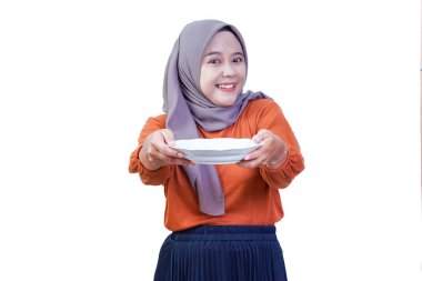 Tesettürlü gülümseyen Asyalı bir kadın iki eli beyaz bir arka planda olan boş beyaz bir tabak sunuyor..