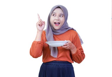Tesettürlü bir kadın elinde tabakla beyaz bir arka planda şaşırmış bir ifadeyle yukarı bakıyor..