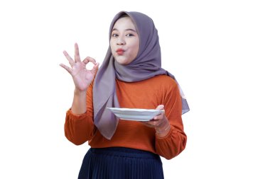 Tesettürlü, neşeli Asyalı bir kadın. Parlak bir şekilde gülümsüyor, tabağı tutuyor ve neşeli bir el hareketi yapıyor..