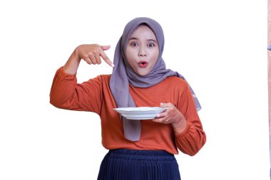 Beyaz bir tabak tutan ve beyaz bir arka planda şaşırmış bir ifadeyle aşağı işaret eden tesettürlü bir kadın..