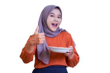 Tesettürlü gülümseyen Asyalı bir kadın boş beyaz bir tabak tutarken başparmağını kaldırıyor. Onaylıyor ve seviniyor..