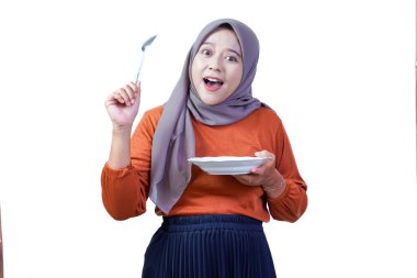 Heyecanlı Asyalı kadın tesettür tabağı ve kaşıkla yukarı bakıyor, neşeli bir ifadeyle, yemeye hazır.