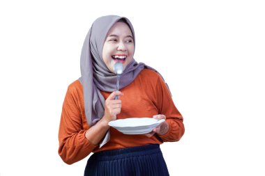 Heyecanlı Asyalı kadın tesettür tabağı ve kaşıkla yukarı bakıyor, neşeli bir ifadeyle, yemeye hazır.