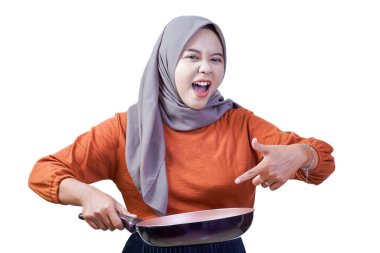 Tesettürlü neşeli Asyalı kadın coşkulu ve neşeli yemek pişirme ruhuyla tavayı işaret ediyor..