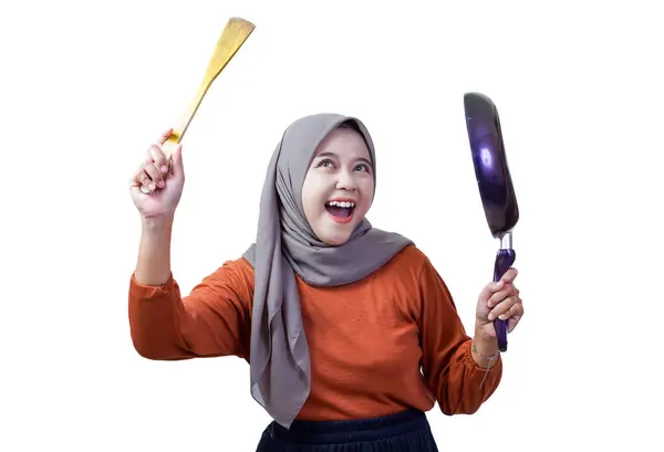 Tesettürlü neşeli Asyalı kadın elinde spatula ve tavayla yemek pişirme pozisyonunda gülümsüyor..