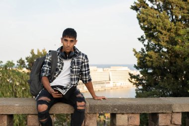 Genç uluslararası öğrenci kampüste çitin üstünde oturmuş kitap okuyor..