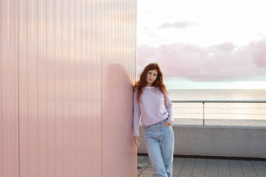 Pembe bir duvara yaslanmış denizin önünde poz veren genç bir kadın. Dramatik gökyüzü.