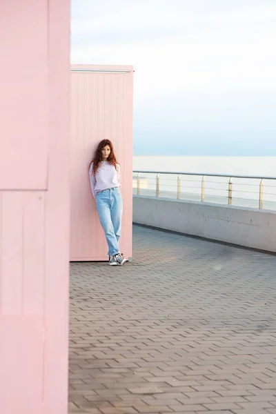 Pembe bir duvara yaslanmış denizin önünde poz veren genç bir kadın. Pastel renklerin paleti.