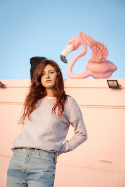 Pembe bir duvarın önünde poz veren genç bir kadın. Arkaplanda flamingo figürü.