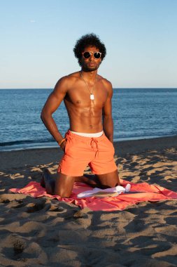 Genç Brezilyalı sporcu, plajdaki bir spor salonunda yoğun bir idman yapıyor, barfiks çekiyor ve parlak turuncu bir mayo giyerken diğer beden eğitimi egzersizlerini yapıyor..