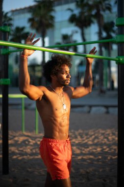 Genç Brezilyalı sporcu, plajdaki bir spor salonunda yoğun bir idman yapıyor, barfiks çekiyor ve parlak turuncu bir mayo giyerken diğer beden eğitimi egzersizlerini yapıyor..