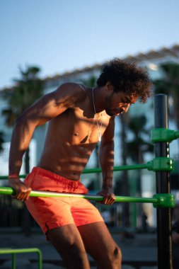 Genç Brezilyalı sporcu, plajdaki bir spor salonunda yoğun bir idman yapıyor, barfiks çekiyor ve parlak turuncu bir mayo giyerken diğer beden eğitimi egzersizlerini yapıyor..