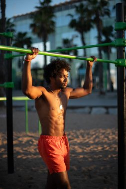 Genç Brezilyalı sporcu, plajdaki bir spor salonunda yoğun bir idman yapıyor, barfiks çekiyor ve parlak turuncu bir mayo giyerken diğer beden eğitimi egzersizlerini yapıyor..