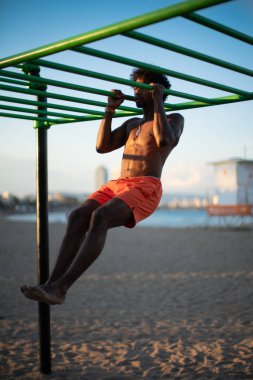 Genç Brezilyalı atlet sahildeki bir spor salonunda yoğun bir idmana katılmış, barfiks çekmiş ve diğer jimnastik egzersizleri yapmış. Turuncu güneşin altında parlak turuncu bir mayo giyerken zayıf ve kaslı vücudunu sergiliyor..