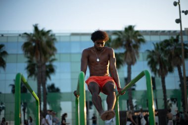 Genç Brezilyalı atlet sahildeki bir spor salonunda yoğun bir idmana katılmış, barfiks çekmiş ve diğer jimnastik egzersizleri yapmış. Turuncu güneşin altında parlak turuncu bir mayo giyerken zayıf ve kaslı vücudunu sergiliyor..