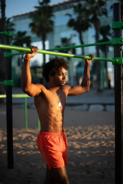 Genç Brezilyalı sporcu, plajdaki bir spor salonunda yoğun bir idman yapıyor, barfiks çekiyor ve parlak turuncu bir mayo giyerken diğer beden eğitimi egzersizlerini yapıyor..