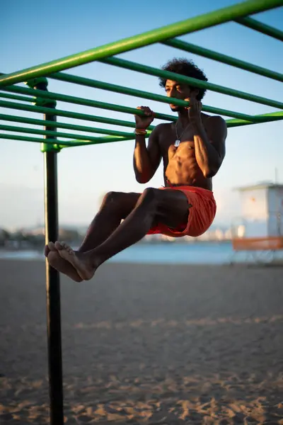 Genç Brezilyalı sporcu, plajdaki bir spor salonunda yoğun bir idman yapıyor, barfiks çekiyor ve parlak turuncu bir mayo giyerken diğer beden eğitimi egzersizlerini yapıyor..