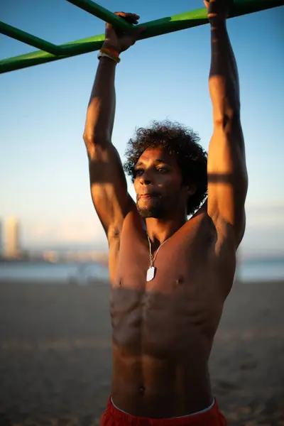 Genç Brezilyalı atlet sahildeki bir spor salonunda yoğun bir idmana katılmış, barfiks çekmiş ve diğer jimnastik egzersizleri yapmış. Turuncu güneşin altında parlak turuncu bir mayo giyerken zayıf ve kaslı vücudunu sergiliyor..