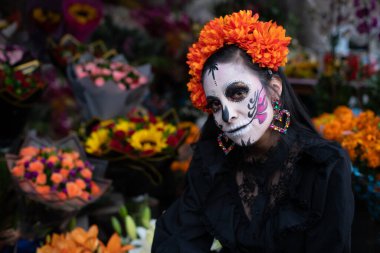 Geleneksel makyajlı bir Meksikalı Catrina, başını şakacı bir şekilde eğmiş, ön planda ve arka planda canlı çiçeklerle çevrili..