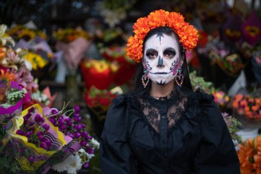 Meksikalı bir Catrina kadife çiçeği tacıyla süslenmiş, kameraya bakıyor, ön planda ve arka planda canlı çiçeklerle çevrili..