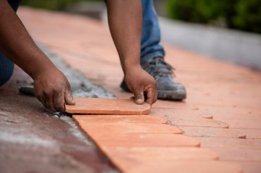 Terracotta çatı fayansını tutan işçilerin yakın çekimi, çatıya yerleştirmek için dikkatlice yerleştiriliyor. Ellere odaklanma, çatı işleminde gerekli olan hassasiyeti ve dikkati gösterir..