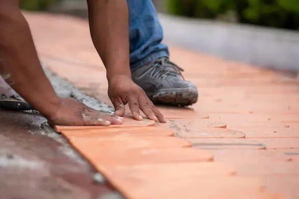 Çimentoya terakotta çatı döşemesi yerleştirmek için tek eliyle vücut ağırlığı uygulayan bir işçinin yakın plan çekimi. Görüntü, seramiğin son konumunu korumak için gösterilen fiziksel çabayı ve hassasiyeti vurguluyor..