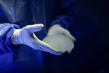 Göğüs implantı tutan beyaz eldivenli bir doktorun ellerine yakın çekim. Görüntü, klinik bir ortamda protezin tıbbi kontrolüne odaklanır..