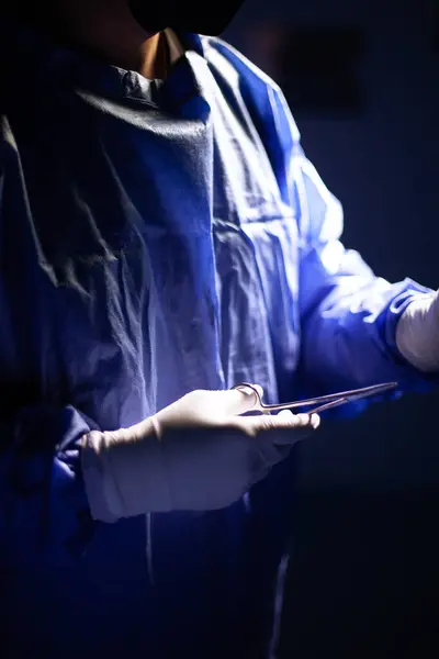 Mavi üniforma giyen bir cerrahın gövde görüntüsü. Eldivenli bir el cerrahi bir makas tutuyor. Alet ve beyaz eldivene odaklan.