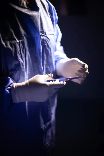 Mavi üniformalı bir cerrahın gövde görüntüsü. Eldivenli bir el cerrahi makası tutarken diğeri dikiş ipliği tutuyor. Tıbbi bir prosedür sırasında bir anı gösteriyor..