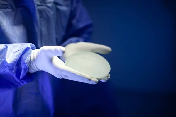 Ameliyat odasında mavi önlük ve beyaz eldiven giyen bir doktor her iki avucunda da şeffaf göğüs implantı tutuyor. Görüntü, bedeni göğsünden kalçasına kadar gösteriyor.