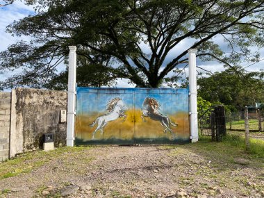 Kırsal bir mülk kapısı arka ayaklarının üzerinde duran iki at heykeliyle süslenir. Sahne kırsal bir girişi gösteriyor.