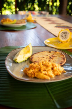 Çırpılmış yumurta, arepa ve avokado içeren tek bir tabak mutfak yüzeyindedir. Arka planda, bir ejderha meyvesi kısmen görünür..