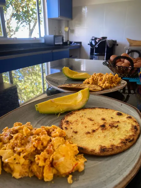 İki tabak çırpılmış yumurta, arepa ve dilimlenmiş avokado bir mutfak yüzeyinde oturur. Düzenli ve rahat, basit bir kahvaltı gösteriyor..