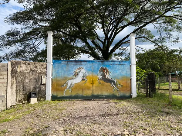 Kırsal bir mülk kapısı arka ayaklarının üzerinde duran iki at heykeliyle süslenir. Sahne kırsal bir girişi gösteriyor.
