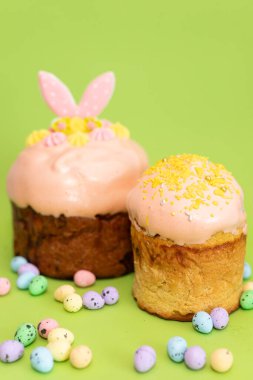 Dikey kompozisyon ve iki geleneksel Paskalya pastası yeşil arka planda yan yana yerleştirilmiş, jöleli ve şenlikli malzemelerle süslenmiş..