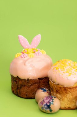 Üzerinde pembe tavşan kulakları ve pastel krema çiçekleri olan Paskalya pastasına yakın plan. Yanında da süslemeli yumurtalar..