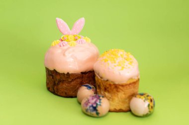 Etrafında dekoratif yumurtalar bulunan parlak yeşil bir yüzeye yerleştirilmiş iki Paskalya pastası neşeli bir tatil kompozisyonu oluşturuyor.