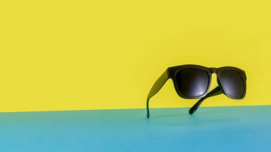 Bir çift siyah güneş gözlüğü parlak mavi ve sarı bir zemin üzerinde süzülüyor gibi görünüyor. Oyuncu kompozisyon ve kalın kontrast, yaz, boş zaman ve minimalizm duygusunu çağrıştırıyor. Kavram için ideal