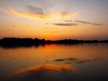 Nehirde güzel bir gün batımı, bulutların yansıması, ufukta ağaçlar, renkli.
