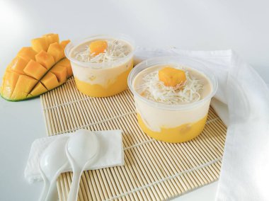 Süt kremalı iki fincan mango tatlısı, bambu soslu rendelenmiş peynir ve beyaz arka planda mango garnitürü..